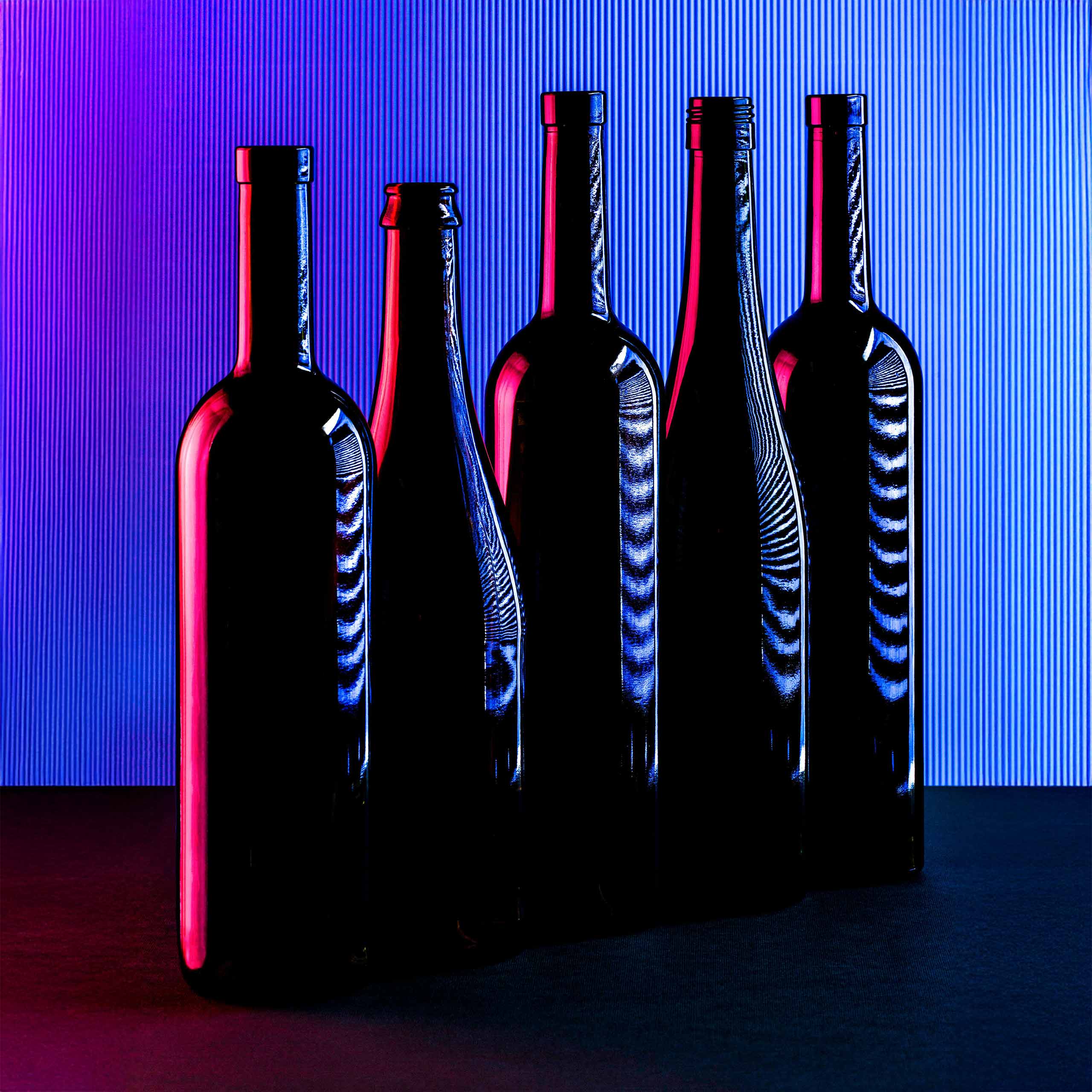 Silhouettes of five tall wine bottles standing side by side against a vibrant backdrop of blue and magenta light, creating reflective highlights and bold color contrasts on the glass.